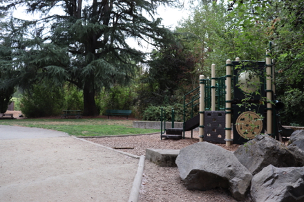 Playground on bark-chip surface – rocks to sit and climb on – bench and picnic table under tree on natural surface